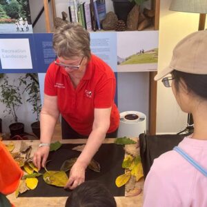 Woman in a red shirt demonstrating to a group of people how to place leaves and twigs artistically on a table.